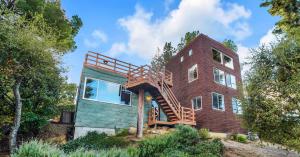 The Benson House by Frank Gehry, a modern two-story home with a wooden staircase connecting two sections, stands on a hillside in Calabasas Highlands, surrounded by greenery and trees under a partly cloudy blue sky.