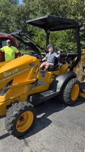 A young child in a blue bucket hat sits in the driver's seat of a large yellow Blue Line Landscape Construction backhoe on a sunny day, holding onto the cab with one hand raised. A man in a neon green shirt stands smiling in the background near a red truc
