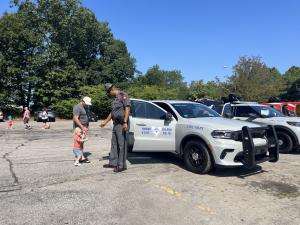 A State Police officer in uniform bends toward a young toddler in a red shirt standing next to a white State Police SUV in a sunny parking lot. An adult in a gray t-shirt and white cap stands beside the child. Additional state police vehicles are visible
