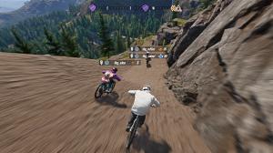 Third-person view of a mountain biker racing against other bikers in mountainous terrain with rocky walls and a dirt track. Motion blur emphasizes the rider's speed as he and the other bikers head toward a dirt ramp.