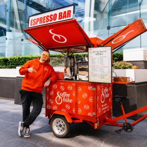 Vladislav Priadko standing beside a red mobile Coffee Bike espresso bar during the 90-day "THE CROSSING – Canada and USA" journey across North America.