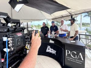 View from behind a television camera looking at an outdoor broadcast set under a canopy tent. Four male commentators wearing headsets stand behind a curved black desk featuring the logos for "The Pro" and "PGA National Resort Palm Beach Gardens."