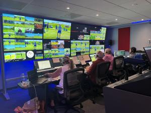 Inside a busy broadcast control room where a crew of four operators sits at desks facing a giant wall of monitors. The screens display multiple live video feeds and camera angles of a golf tournament, while the crew members monitor laptops and production