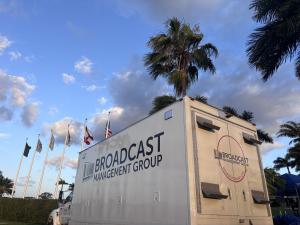Exterior shot of a large white broadcast production truck with the "Broadcast Management Group" logo printed on its side and back. The truck is parked outdoors against a backdrop of palm trees and a line of flags under a partly cloudy blue sky.