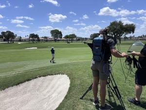 A camera operator, wearing a blue LiveU backpack unit, films a golfer on a bright, sunny day at a golf course. A sand bunker is visible in the immediate foreground, and manicured green fairways stretch out toward houses and trees under a blue sky with flu