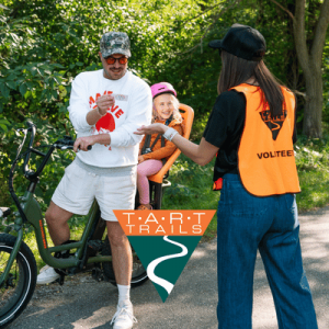 Man on bike wearing sunglasses interacts with a volunteer in an orange vest, while a child in a helmet smiles in a green outdoor setting.