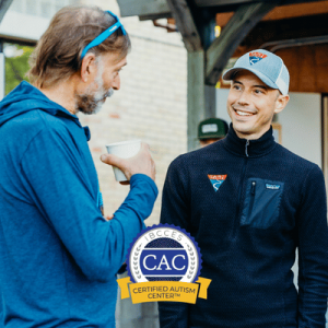Two men smiling and chatting, one holding a cup, with a Certified Autism Center (CAC) logo overlay.
