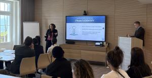 Dr. Julie Meltzer stands and speaks in front of a presentation screen reading "The Solutionary Framework, Phase 1: IDENTIFY," with a seated audience in the foreground and a colleague at a podium in the background.