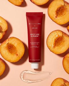 A tube of Joylux x Respin Juicy Like a Peach™ moisture-rich vaginal gel is displayed on a peach-colored background surrounded by fresh peach halves, with a clear gel swatch beneath the product.