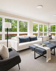 Bright professionally staged living room featuring a white sofa, modern accent chairs, oversized windows, and contemporary furnishings overlooking wooded views