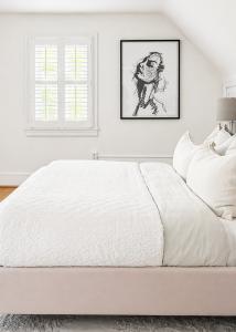 Professionally staged primary bedroom with upholstered bed, layered white bedding, plantation shutters, contemporary artwork, and neutral designer décor.