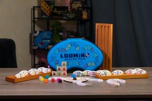 Loomino curved-tile domino game by Queensell, with a blue carrying case, colored curved tiles in wooden racks, train tokens, and train-symbol dice on a wooden table.