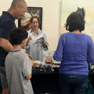 Four people interact at a stall with jars labeled "blood," "frogs," and "lice," featuring a "Witches Market" sign and horse paintings in the background.