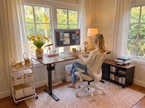 Woman working at an OffiGo standing desk in a bright home office with a monitor, ergonomic chair, printer, and mobile file cabinet.