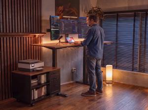 Man standing at an OffiGo height-adjustable desk with multiple monitors in a dark home office, next to a mobile printer and file storage cabinet.