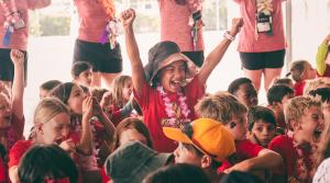 A cheering child in a red Kidventure shirt raises both arms while surrounded by a large group of campers during All Camps Day in Houston.