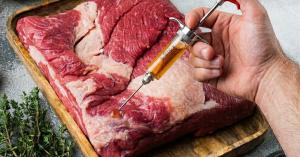 A man's hand using a SpitJack SQUIRT 50 meat injector to inject seasoned brine into a trimmed raw brisket on a wooden board.