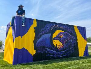 Blue Robin Seated on a Shipping Container Featuring His Painted Crow Mural
