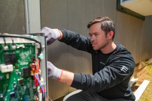 A technician wearing a long-sleeved black shirt and protective work gloves carefully works on an open electrical control board of an outdoor heat pump against a house.