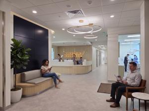 Reception area of Austin Institute of Dental Medicine with curved seating, wood finishes, and modern ring lights. Patients wait while staff assist at the front desk, and dental professionals treat a patient in a bright exam room in the background.