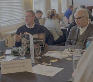 Two participants from the TeachingAbout.AI Convening sit at a table with other educators discussing the Rochester Provocation "Assessments were a problem before AI."