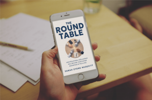 A hand holding a mobile phone with an image of a book called The Round Table on it, hovering above a wooden table