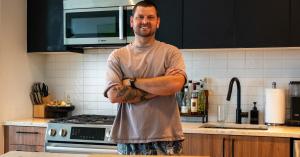 A portrait of Kyle Taylor standing with arms crossed in a modern kitchen with black cabinets, stainless steel appliances, and cooking ingredients neatly arranged on the countertop.