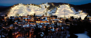 Night view of a snow-covered ski resort with illuminated slopes and a village below, surrounded by dark wooded hills.