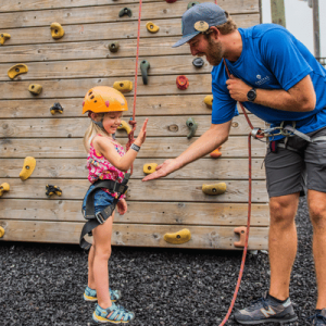 A girl in climbing gear high-fives an instructor in front of a rock climbing wall.