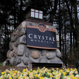 Stone monument with a lantern top and "Crystal Mountain" sign, surrounded by yellow daffodils and tall trees in the background.