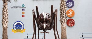 Sculpture of a large spider between two palm trees at the Disease Vector Education Center, featuring insect icons and certification logos.