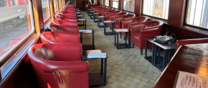Interior of a vintage train car with rows of red leather armchairs and small black tables beside large windows.