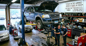 Two mechanics in a busy auto repair shop work underneath a rusty blue Ford SUV raised on a hydraulic lift. The vehicle's hood is open, and the mechanics are servicing a transmission component supported by a jack. The background features tires stacked on s