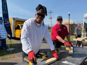 Ji'Ayir Brown, wearing sunglasses and a white long-sleeve shirt, and Jordan Allen, in a red SHP polo and cap, use orbital sanders on wood boards at an outdoor bed build event.