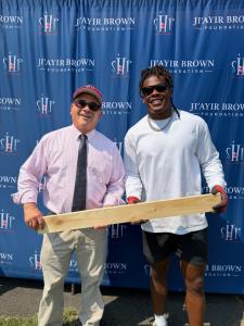 Trenton Mayor Reed Gusciora, in a pink dress shirt and tie, and Ji'Ayir Brown, in a white shirt and sunglasses, hold a wooden board together while posing in front of an SHP and Ji'Ayir Brown Foundation step-and-repeat backdrop.