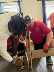 Ji'Ayir Brown, wearing a white long-sleeve shirt, uses a power drill on a wooden bed frame while Jordan Allen, in a red SHP polo and cap, holds the frame steady inside a bedroom.