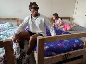 Ji'Ayir Brown, wearing sunglasses and a white long-sleeve shirt, laughs while sitting on a new bed with a unicorn comforter, as a young girl in pink pajamas smiles beside him.