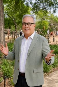 Rich DePalma, Executive Director of the Texas Recreation and Park Society, stands outdoors in a park wearing glasses, a light green blazer, and a white shirt, smiling with his hands open in a welcoming gesture.