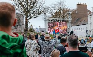 Crowds gather in an Isle of Man town square to watch King Charles III’s coronation on a large outdoor screen, with a child in green in the foreground.
