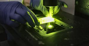 Gloved hands positioning a sample on an illuminated microscope stage in a laboratory.