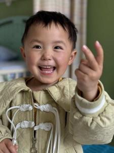 Four-year-old Hao smiles and holds up two fingers while wearing a light green quilted jacket.