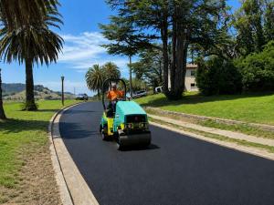 Worker in orange high-visibility shirt operates a teal and yellow double-drum vibratory asphalt roller on a freshly paved palm-lined roadway. Rolling hills, Mediterranean-style home and clear blue sky in the background.