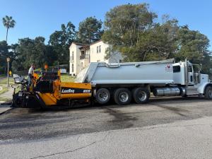 Yellow LeeBoy 8520 asphalt paver positioned in front of a white SBS Roadlines Inc. dump truck on a residential street. Operator stands at the paver controls. Mission Revival two-story building with red tile roof and palm tree behind.
