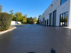 Newly paved two-lane roadway with a bright double-yellow center stripe and a raised speed bump marked with fresh white striping in the foreground. Mission Revival buildings with red tile roofs on the right, mature trees on the left and clear blue sky abov
