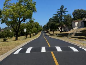Freshly paved apartment complex parking lot with crisp white parking stall striping and smooth dark asphalt surface. Landscaped shrubs, mature trees and a residential apartment building visible in the background under clear blue sky. Utility transmission 