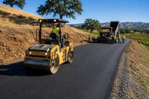 Yellow asphalt roller compacts a freshly paved road as a paving crew works beside a vineyard in California’s North Bay.