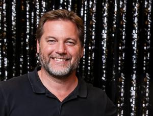 Smiling man with short brown hair and a beard, wearing a black polo shirt against a dark, reflective background.
