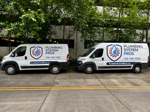 Two white Plumbing System Pros service vans parked side by side on a Houston street in front of a commercial building. The branded service vehicles display the company logo, website, and phone number, representing residential and commercial plumbing servi