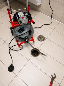 Professional drain cleaning machine with a sewer cable inserted into a commercial floor drain during a drain cleaning service. The plumbing equipment is positioned on a tiled restroom floor, demonstrating routine maintenance to remove drain blockages and