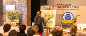 Artist painting floral artwork on easel while audience watches, with St. Johns Cultural Council and Certified Autism Center logos displayed.